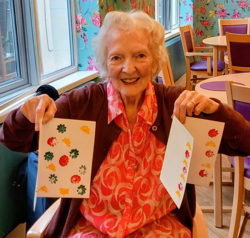 An elderly woman sits in a brightly decorated room, smiling and holding up two sheets of paper with colourful leaf prints, celebrating creativity.