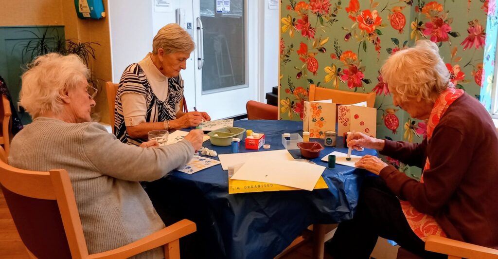 Three elderly women sit around a table enjoying colourful arts and crafts, painting and assembling items as lively conversation fills the brightly decorated room adorned with floral wallpaper.