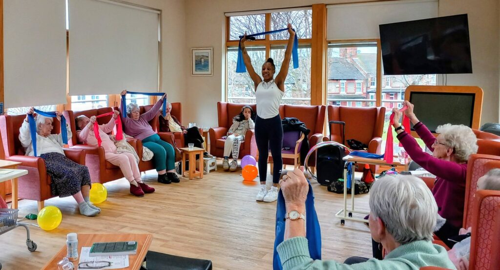 A group of elderly people in a common room enjoy a lovely afternoon, taking part in a seated exercise class, following a standing instructor and using resistance bands.