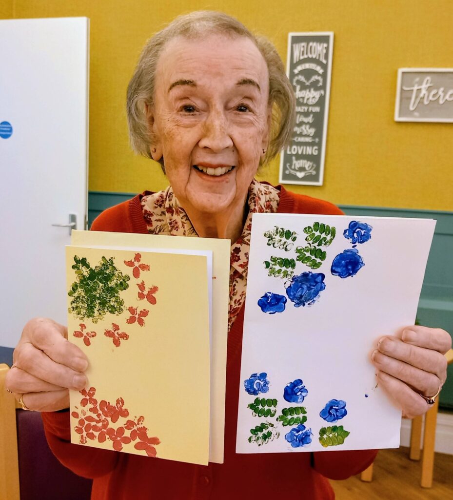 Elderly woman smiling and holding two handmade cards, displaying creativity and colour with vibrant red and blue floral prints.