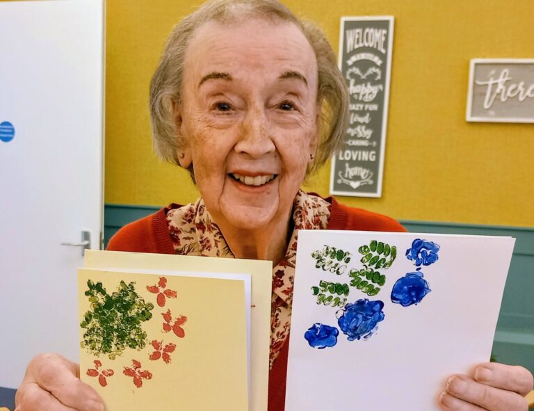 Elderly woman smiling and holding two handmade cards, displaying creativity and colour with vibrant red and blue floral prints.