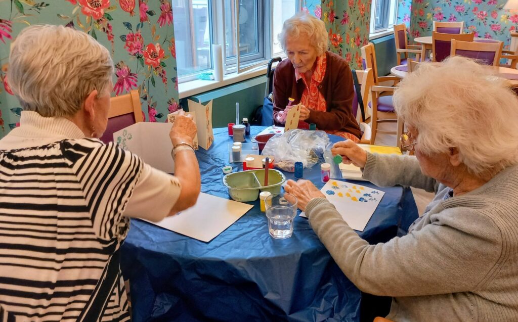 Three elderly women sit at a table covered with a blue cloth, chatting and being creative as they paint and make crafts using paper, paint bottles, and brushes in a bright room adorned with floral decorations.