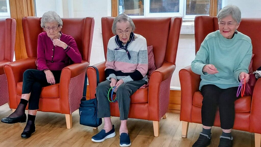 Three elderly women, simply the best at afternoon fun, sit in armchairs playing a hoopla game, aiming rings at colourful cones arranged on a wooden floor.