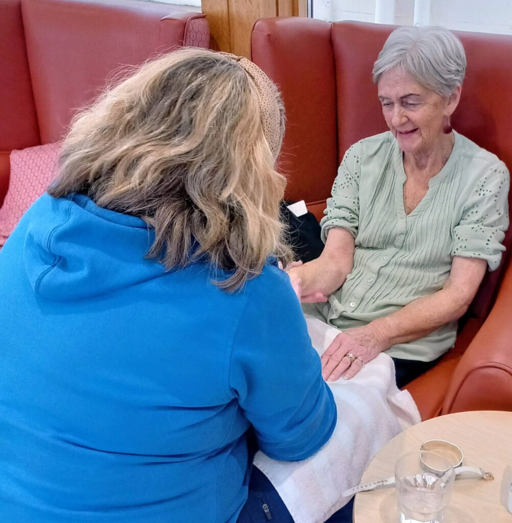 An older woman sits in an armchair smiling as another person in a blue top holds her hand, possibly giving her a manicure. A glass of water sits on the nearby table.