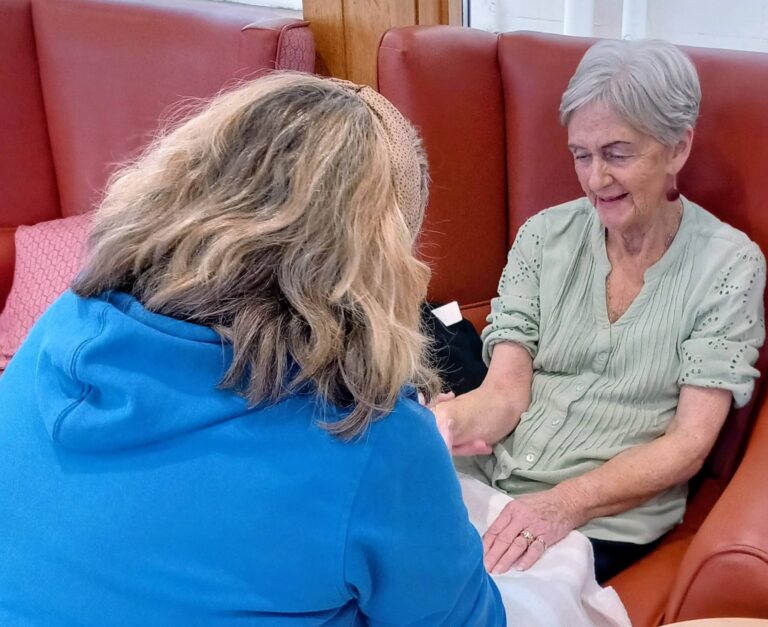 An older woman sits in an armchair smiling as another person in a blue top holds her hand, possibly giving her a manicure. A glass of water sits on the nearby table.