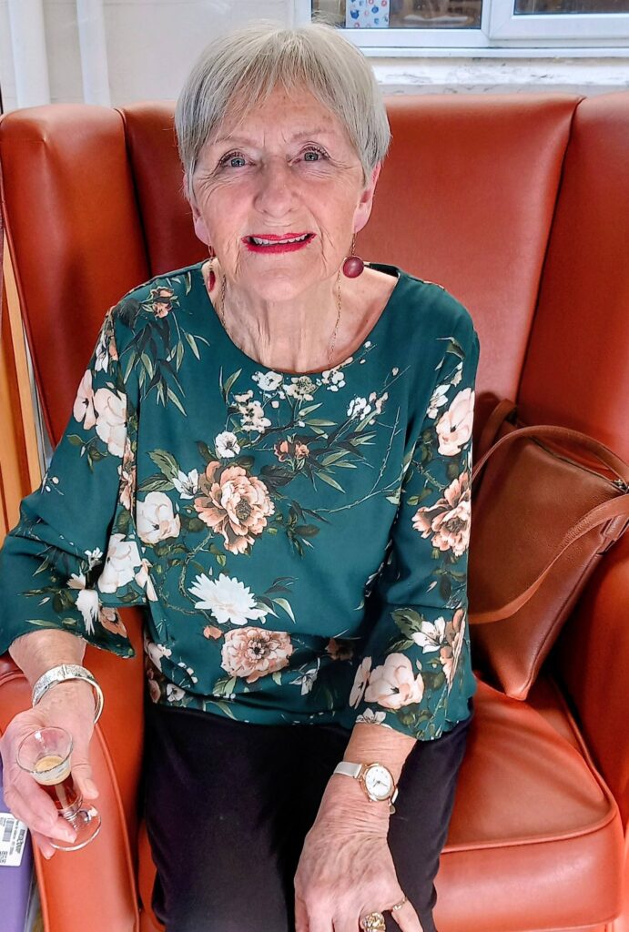 An older woman with short grey hair sits on an orange armchair, wearing a green floral top, holding a small glass, and smiling at the camera. A brown bag rests beside her.