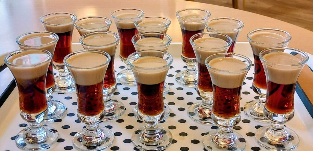 A tray holding several shot glasses filled with layered brown and cream-coloured alcoholic drinks, all arranged in tidy rows.