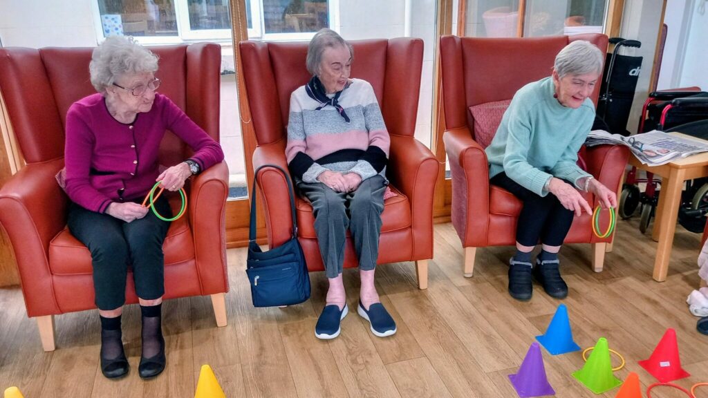On a cheerful afternoon, three elderly women seated in armchairs play a hoopla game, tossing plastic rings at colourful cones—truly the loveliest way to enjoy one another’s company.