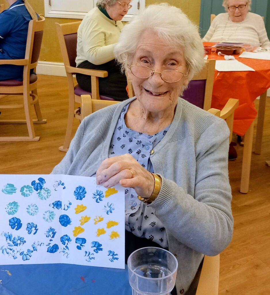 An elderly woman seated at a table holds up her colourful artwork depicting blue and yellow flowers, smiling at the camera. Other pensioners are engaged in lively conversation in the background.