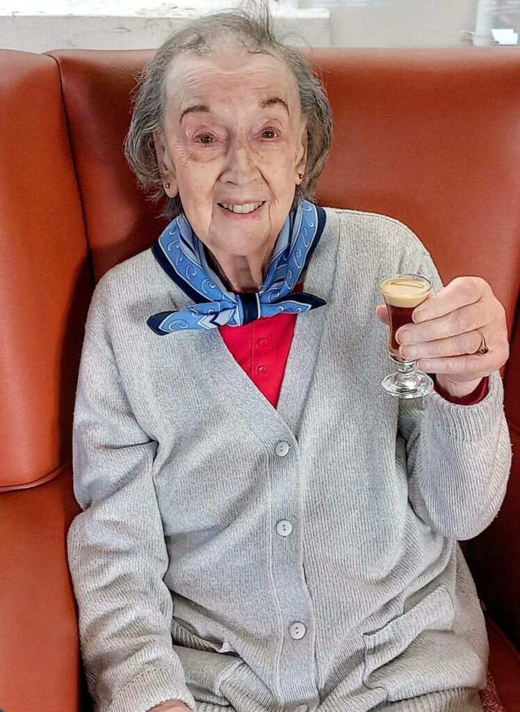 Elderly woman sits on an orange chair, smiling and holding a small glass of layered drink whilst wearing a grey cardigan, red top, and blue scarf.