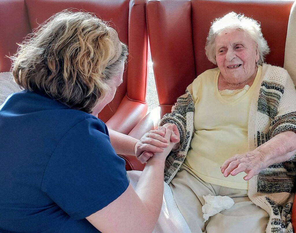 A carer in blue scrubs holds hands with an elderly woman seated on a chair, both smiling and chatting together.