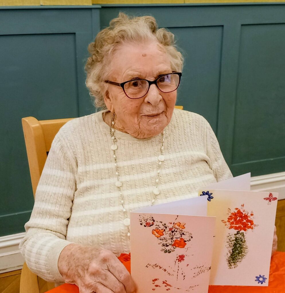 An elderly woman with glasses sits at a table, holding two colourful greeting cards decorated with floral designs. She is wearing a cream-coloured jumper and a beaded necklace.