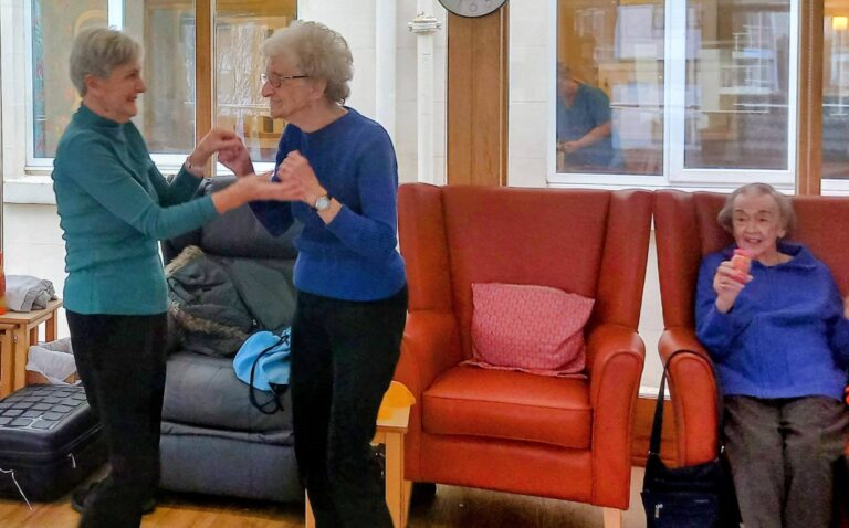 Two elderly women are dancing to music in a sitting room while another elderly woman, seated in a red armchair, smiles and holds a small object.