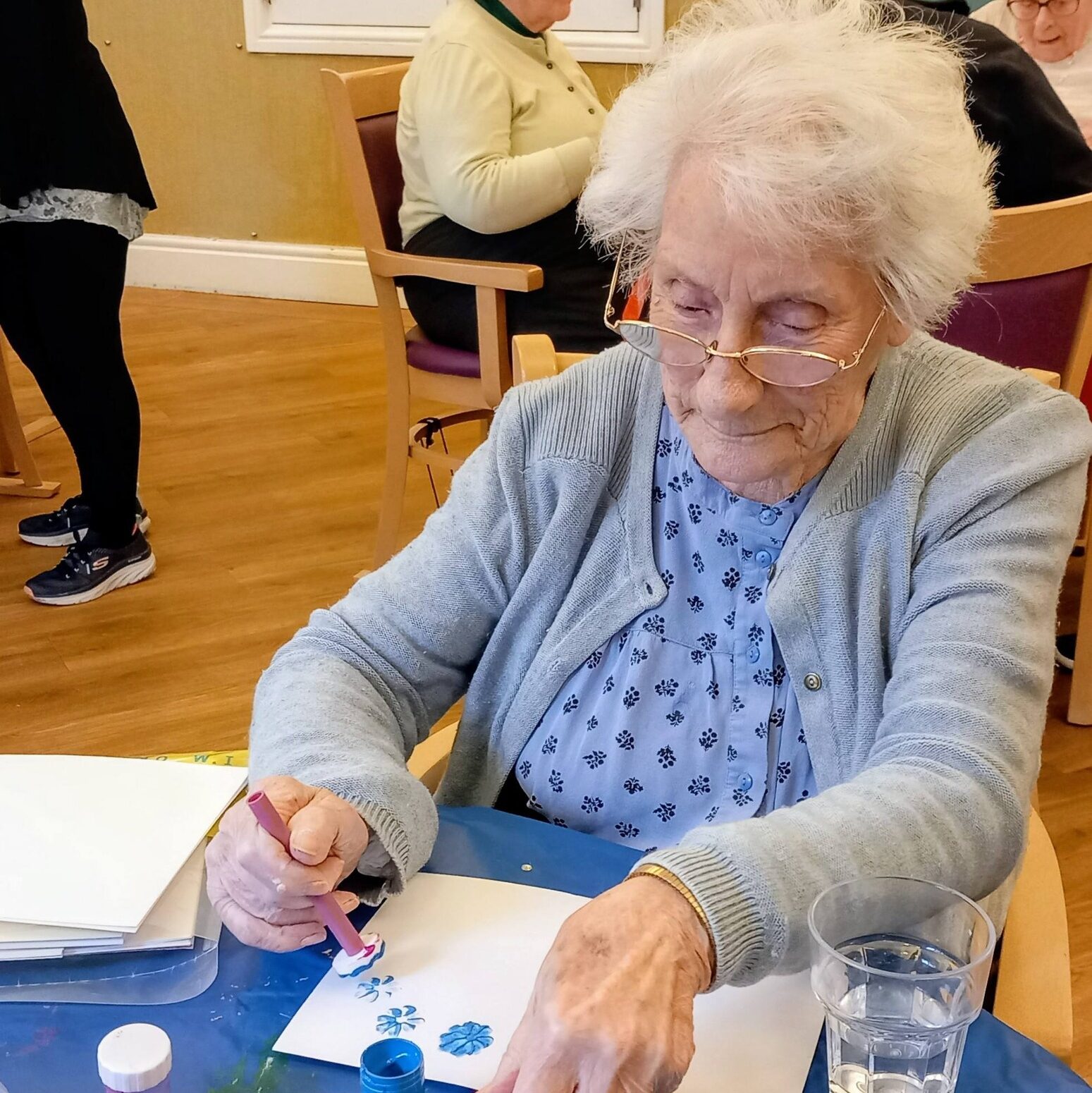 Elderly woman seated at a table painting with blue paint, surrounded by art supplies and a glass of water. Creativity flows as she enjoys her artwork, whilst conversation murmurs in the background.