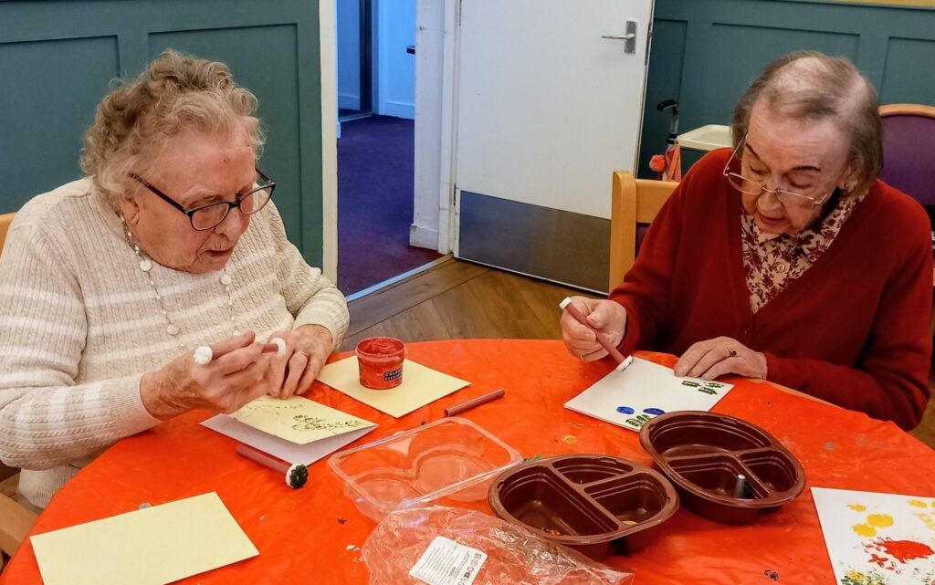 Two elderly women sit at a table, chatting as they decorate colourful cards with felt-tip pens and paint. Arts and crafts supplies are strewn across the orange tablecloth before them.