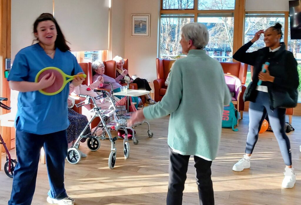 A nurse holding a hoopla game stands with an elderly woman on a cheerful afternoon, while others sit and watch in a sunlit communal room at Simply the Best care home.
