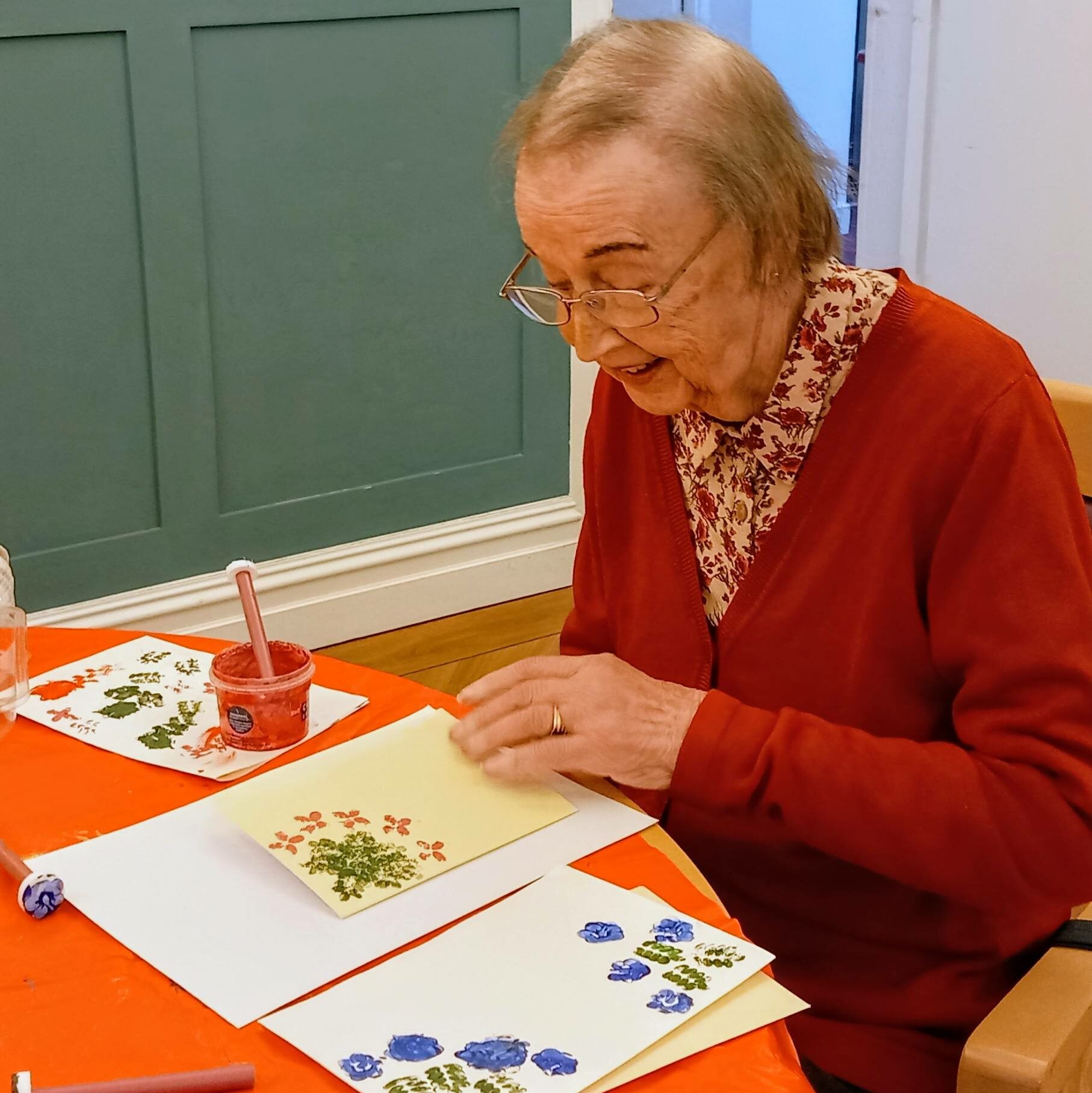 An elderly woman sits at a table, expressing her creativity as she creates colourful floral art on paper using paints and stamps. Both blank and decorated sheets are spread out before her.