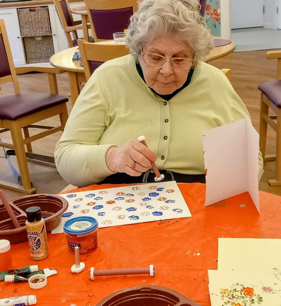 An elderly woman sits at a table, fully absorbed in creativity as she paints colourful flower patterns on paper, surrounded by art supplies in a well-lit room.