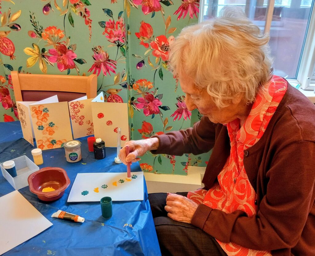 An elderly woman sits at a table, using stamp tools to paint colourful flower patterns. Her creativity shines amid art supplies and finished work on a blue tablecloth, with floral wallpaper in the background.
