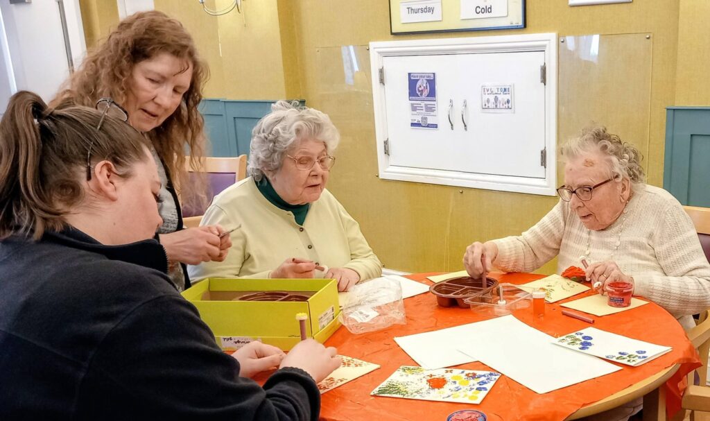 Three elderly women and two carers sit around a table, taking part in a colourful craft activity that encourages creativity and conversation in a bright, well-lit indoor setting.