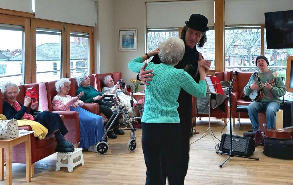A group of elderly people sit and watch as a man in a top hat dances with a woman; a musician plays the guitar and sings, filling the room with rhythm and fun.