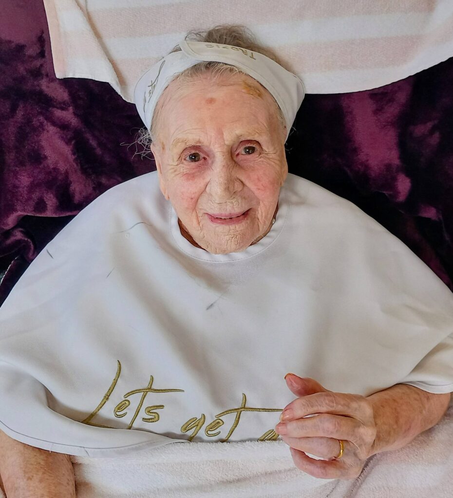 Elderly woman wearing a white headscarf and a draped white cloth featuring gold lettering, seated and looking at the camera, with a purple blanket in the background.
