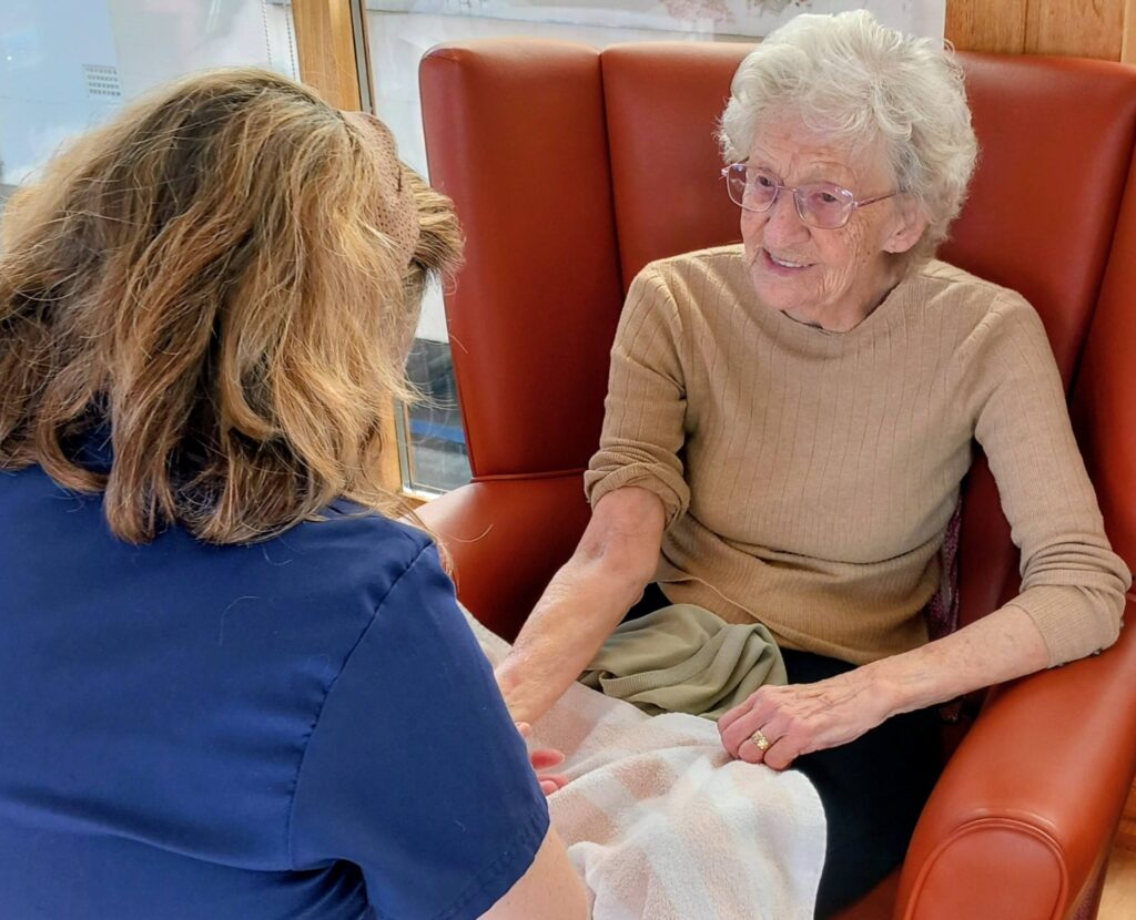 An elderly woman sitting in a red armchair smiles as she holds hands with another person dressed in a blue shirt.