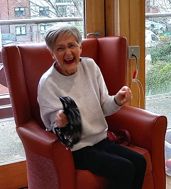 An elderly woman sits in a red armchair, smiling as she plays a black tambourine, adding fun and rhythm to the sunlit room with large windows.