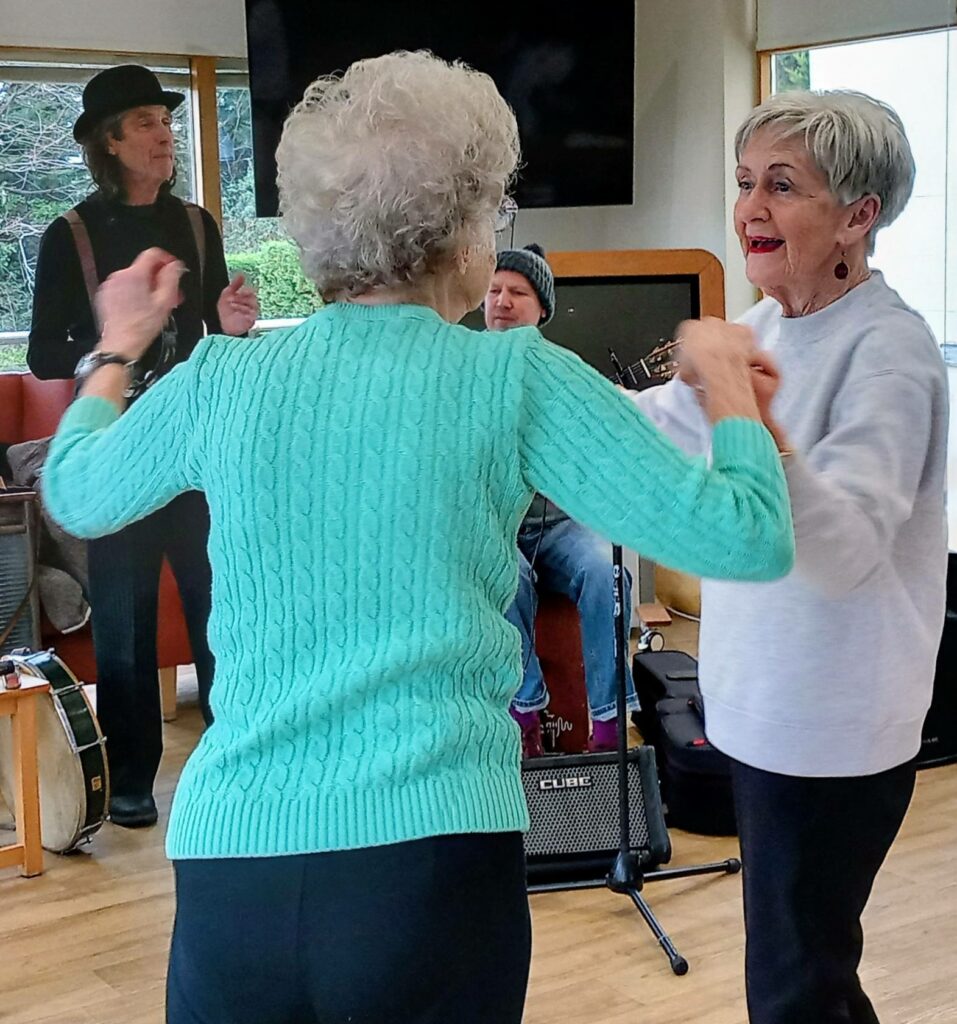 Two older women dance together indoors, moving with enjoyment and rhythm whilst musicians perform in the background.