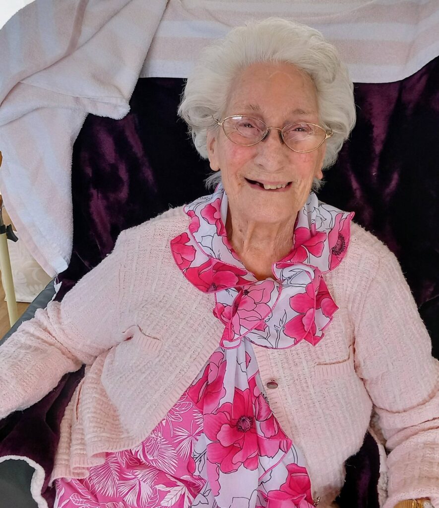 Elderly woman with white hair and glasses, dressed in a pink floral dress and light pink cardigan, seated and smiling at the camera.