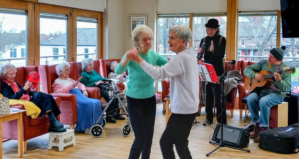 Two elderly women dance together in a communal lounge, moving with lively rhythm as other pensioners look on. A small band featuring a guitarist and clarinettist adds to the cheerful atmosphere in the background.