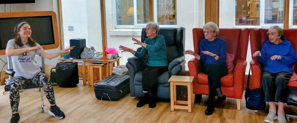 Four elderly women in armchairs and a young woman on a chair raise their arms, exercising together with cheerful smiles in a brightly lit room.