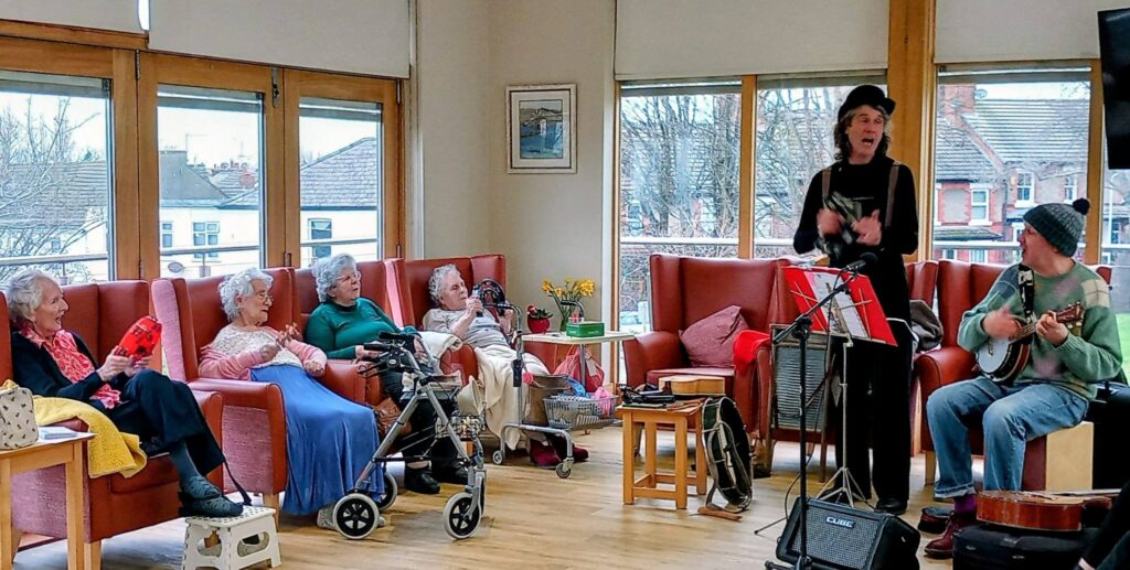 A small group of elderly people sit in armchairs, enjoying the lively rhythm as two musicians perform in a bright communal room with large windows.