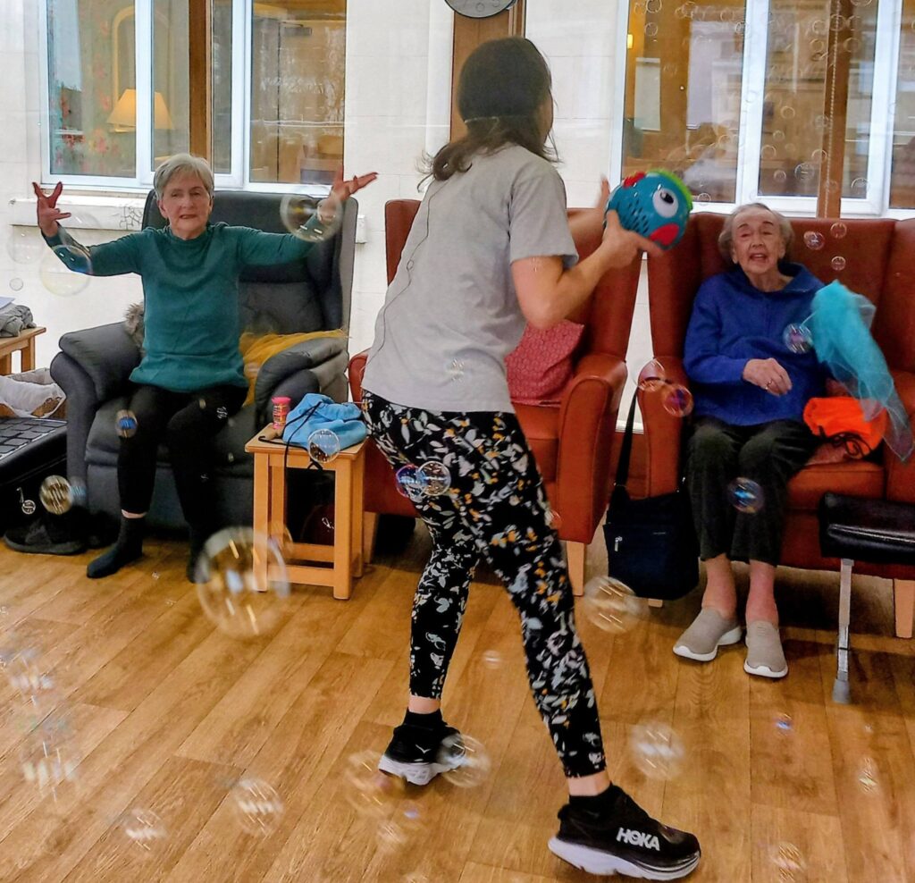 A young woman stands amid soap bubbles, holding a puppet, while gentle music plays. Two elderly women sit in armchairs, smiles lighting up their faces as they reach out towards the playful bubbles.