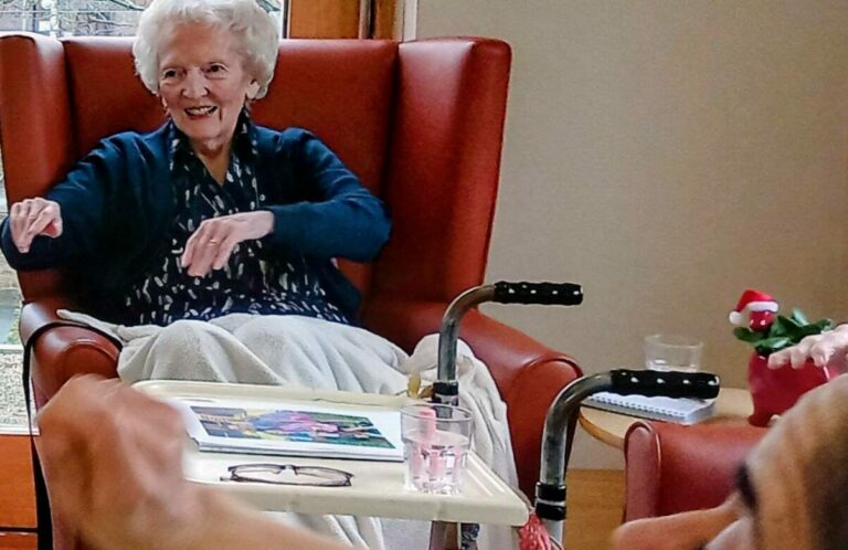 An elderly woman sits in a red armchair, smiling and gesturing as another woman lifts her arm in the foreground. A walking frame and tray with items are by the chair, capturing a warm, music-filled moment.