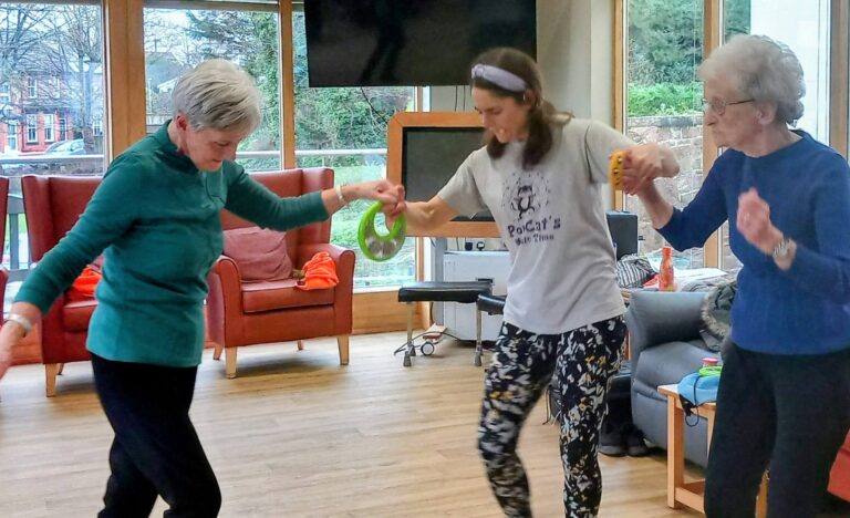 Three women, all smiling, hold hands and dance to music in a bright room with large windows, wooden flooring, and orange chairs in the background.