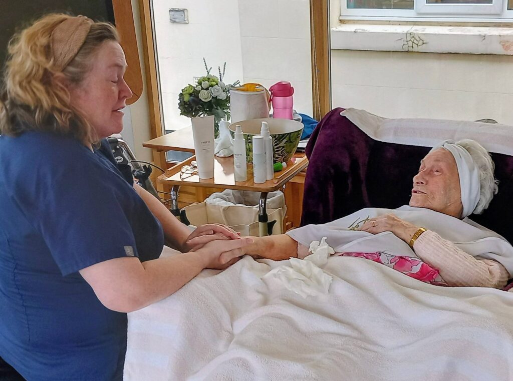 A carer holds the hand of an elderly woman lying in bed, covered with a blanket, in a bright room with personal care items and flowers close by.