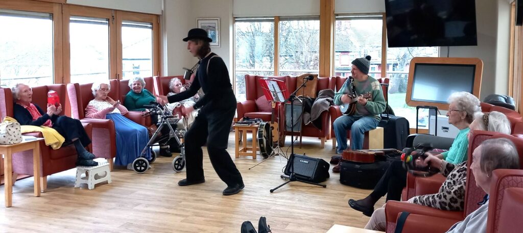 A group of elderly people sit in a bright room, enjoying the fun and rhythm as a performer in a hat and braces entertains them, whilst another plays stringed instruments.