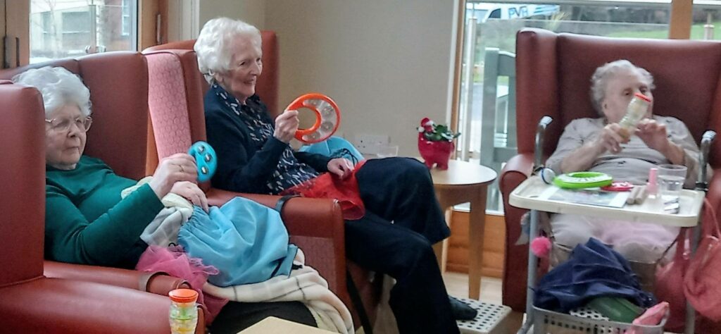 Three elderly women sit in armchairs at a care home, holding colourful musical instruments, their smiles revealing how much they enjoy this uplifting, music-filled indoor activity.