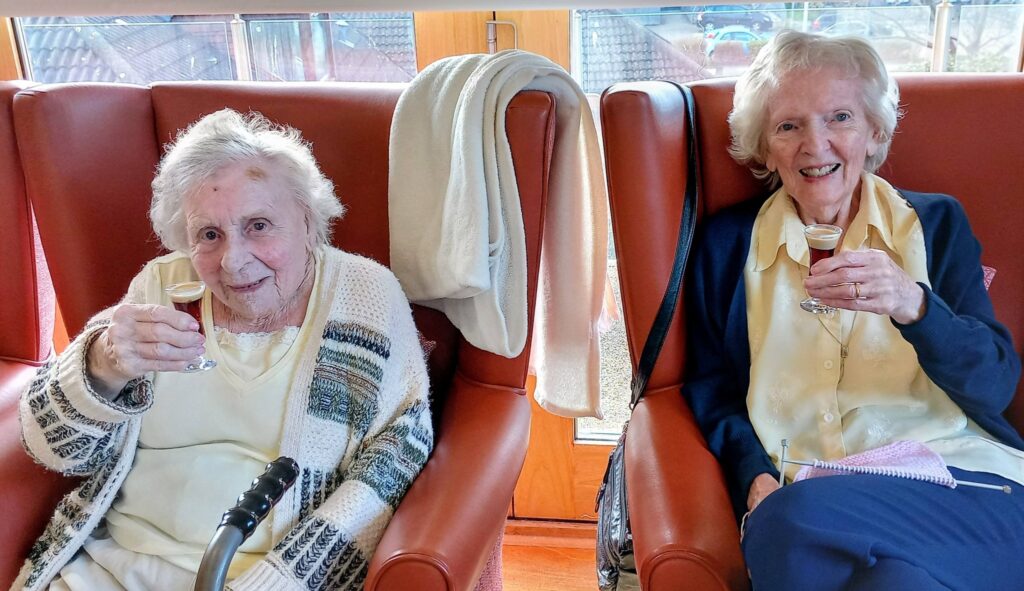 Two elderly women sit in red chairs, each holding a small glass containing a brown drink, smiling at the camera. A white towel is draped over the back of one chair.
