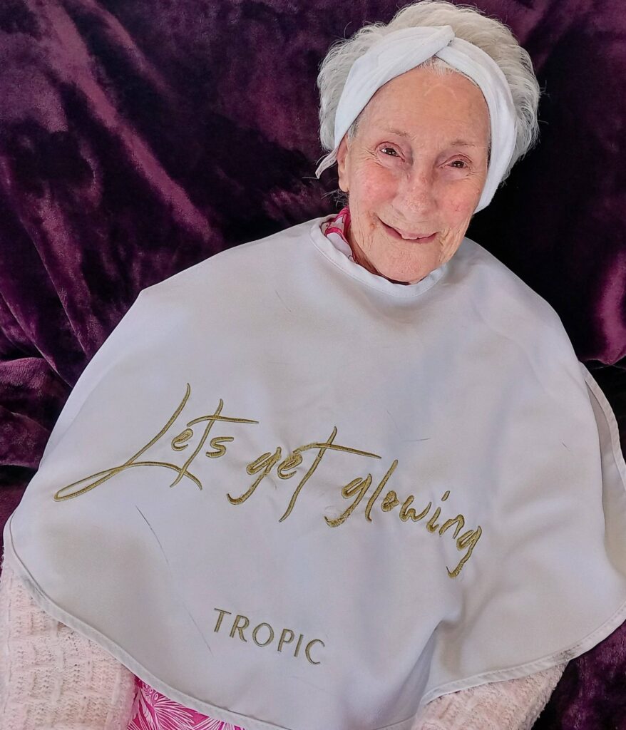 Elderly woman smiling, wearing a white headband and a beauty cape with the words 'Let's get glowing' and 'TROPIC' embroidered in gold.