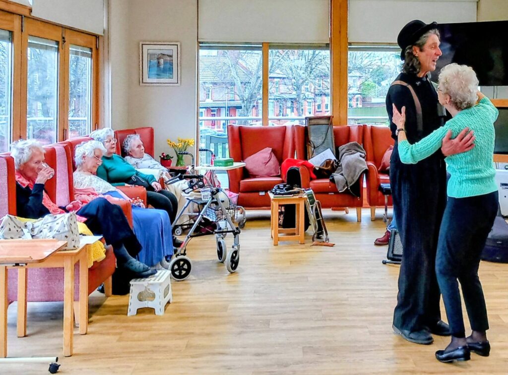 A performer in fancy dress dances with an elderly woman to a lively rhythm, while others sit and watch in a brightly lit common room with large windows and red chairs.