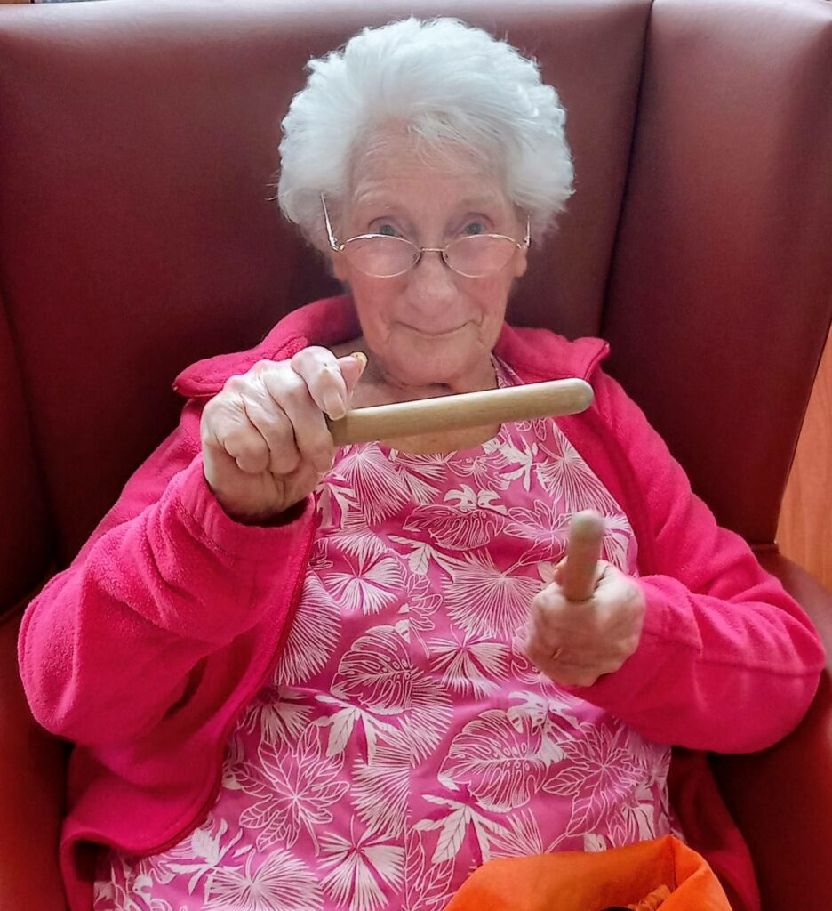 Elderly woman with white hair and glasses, dressed in a pink patterned top and jacket, seated in an armchair holding two wooden percussion sticks, smiles as she enjoys making music.