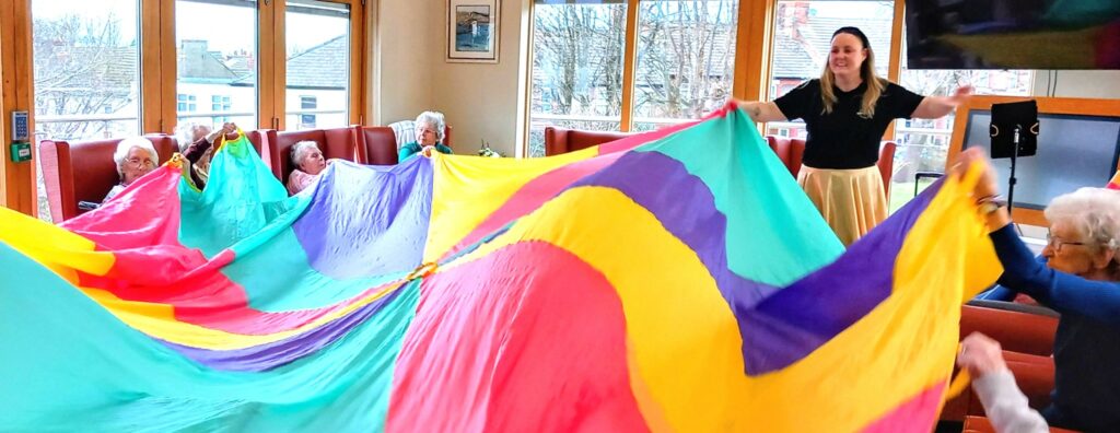 A group of elderly people and a carer enjoy an Olympic-themed session, holding and waving a large, colourful parachute in a bright room with large windows.