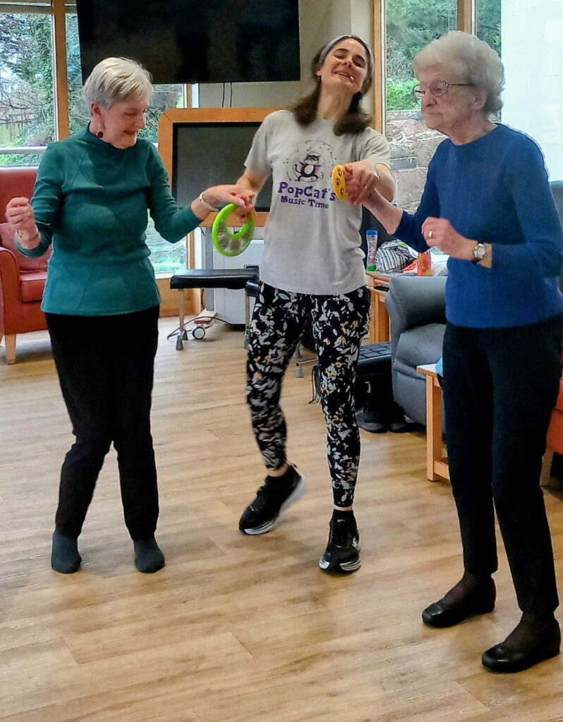 Three women—two elderly and one younger—stand indoors with bright smiles, holding tambourines as they take part in a joyful music or dance activity in a well-lit room.