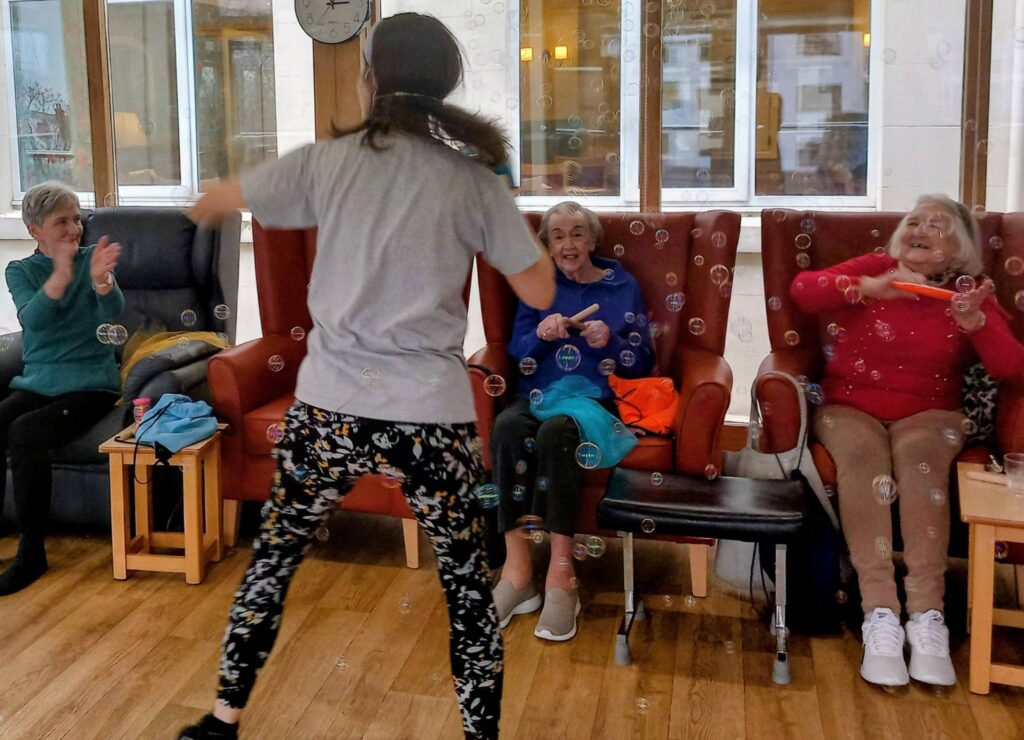 A woman stands facing three elderly women seated in armchairs, filling the brightly lit room with bubbles and smiles as they all share a joyful moment together.