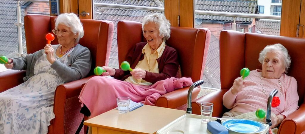 Three elderly women sit in armchairs, shaking colourful maracas during a lively music and movement session. Glasses of water sit on a table before them in a bright room with large windows.