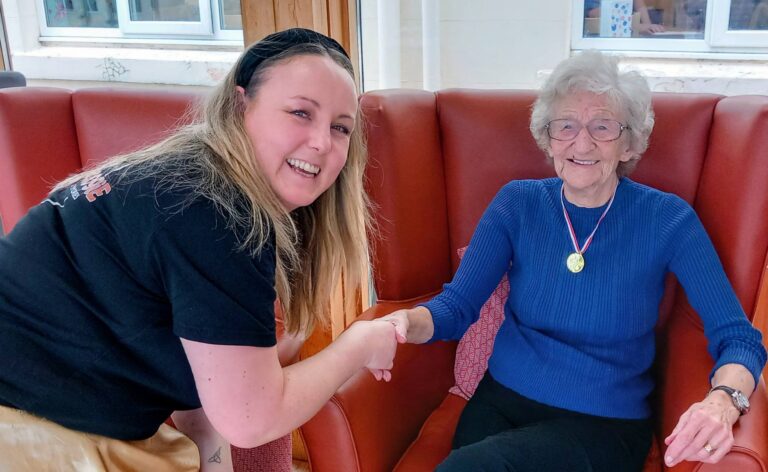A younger woman shakes hands with an older woman seated in a red chair, who is smiling and wearing a gold medal round her neck following an Olympic-themed session.