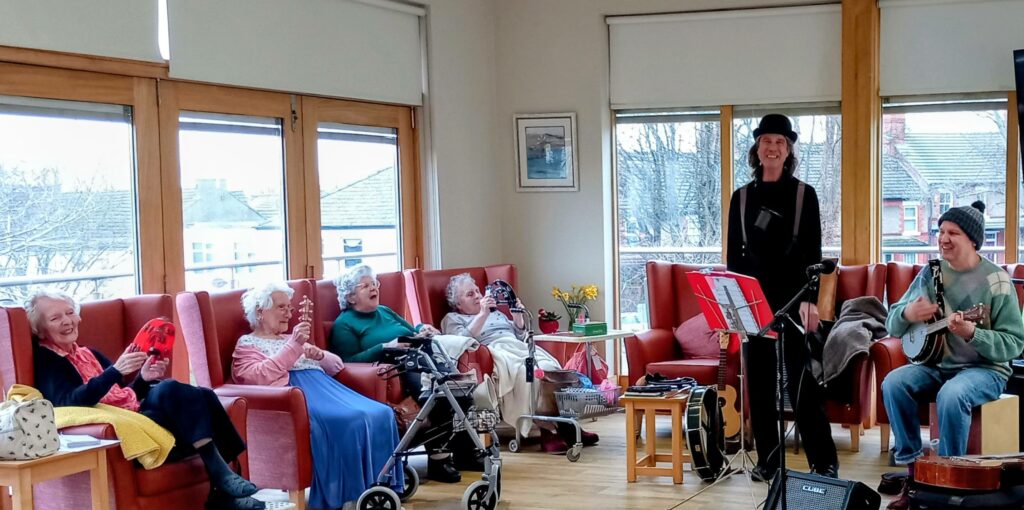 Two musicians bring rhythm and fun to a group of elderly women seated in a bright room with large windows; some are clapping and smiling, while others use walking frames.