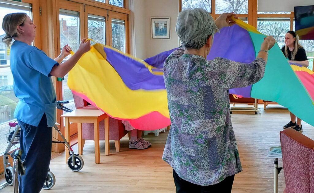 Three women hold and lift a colourful parachute during a music and movement session in a bright room with wooden floors and large windows; a walking frame and chairs are visible in the background.